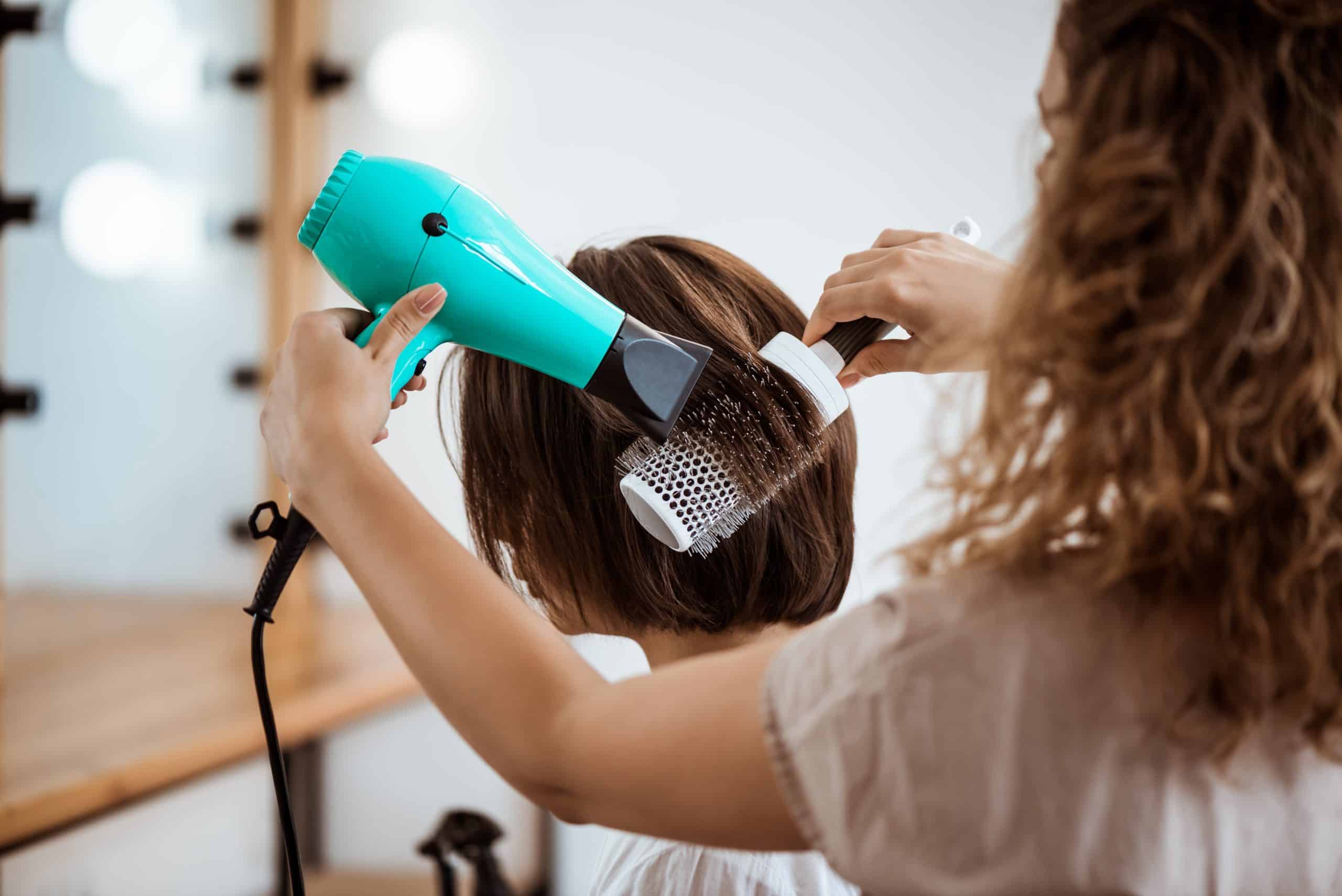 A hairstylist blow-drying a client's short brown hair using a round brush and a teal hairdryer in a salon setting.