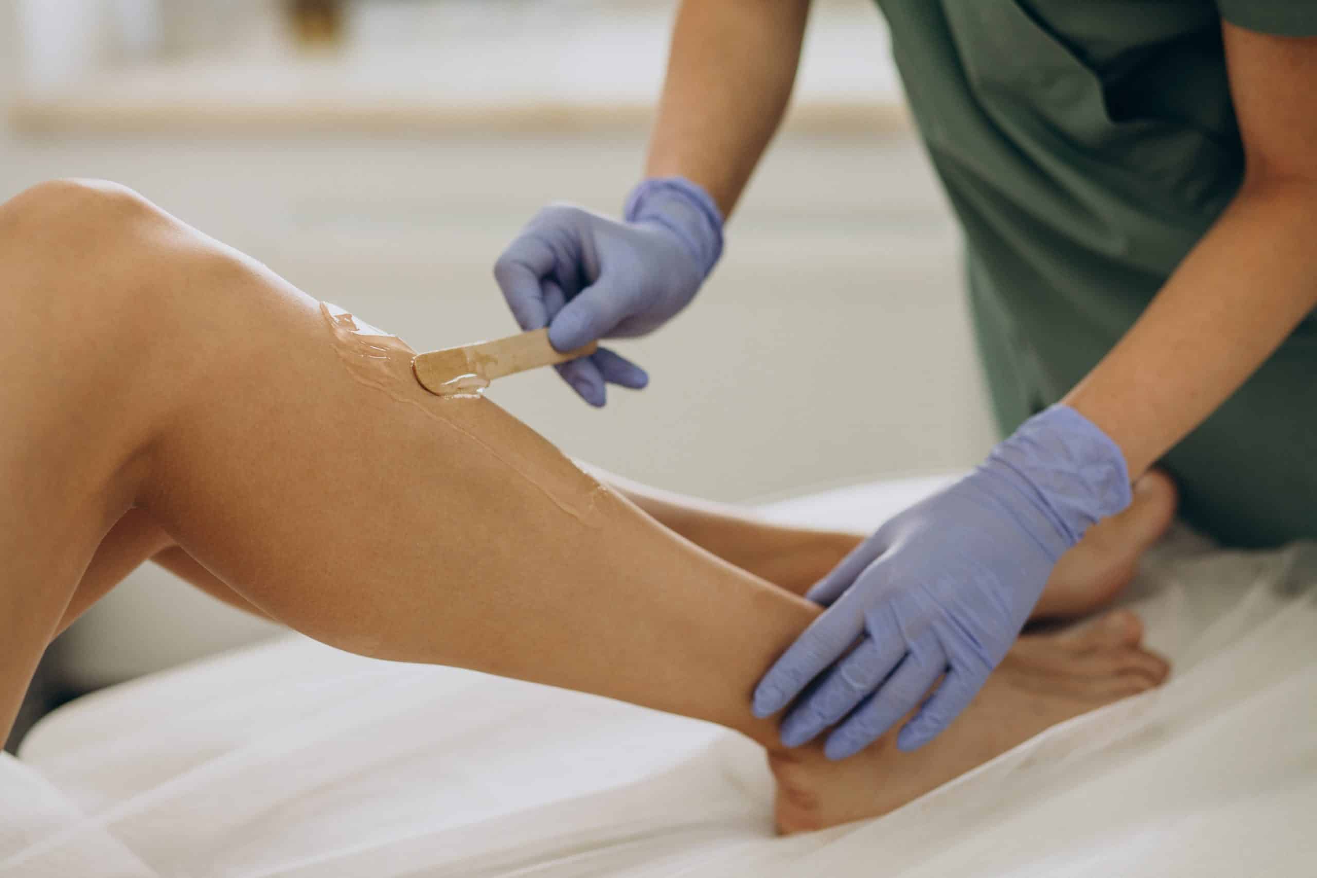 A person wearing purple gloves applies wax to another person's lower leg with a wooden spatula, preparing for a hair removal treatment in a spa or salon setting.