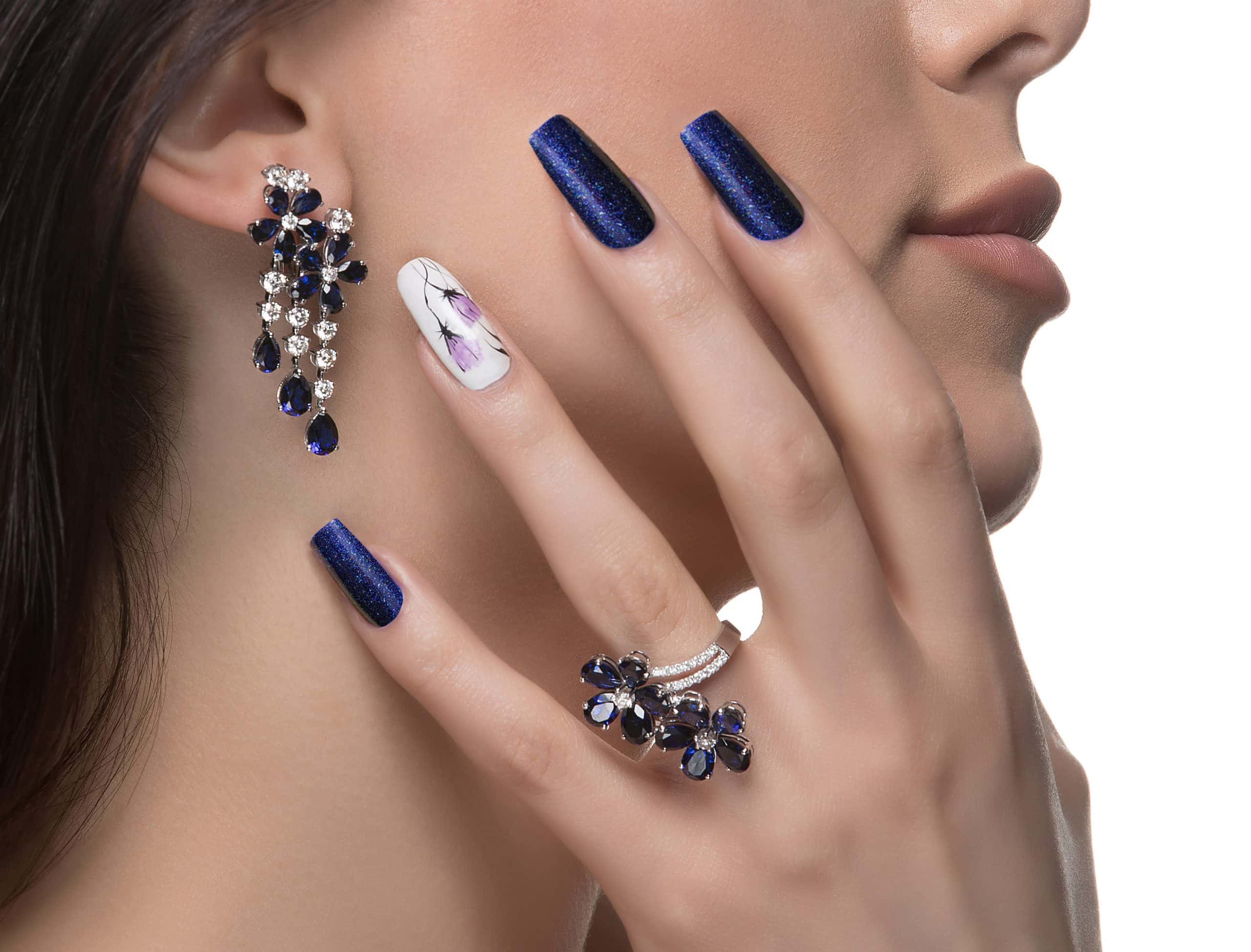 A close-up of a woman’s face and hand showing blue gemstone earrings and ring, with long blue nails. One nail is white with a purple floral design. The woman’s skin is smooth and her lips are slightly parted.